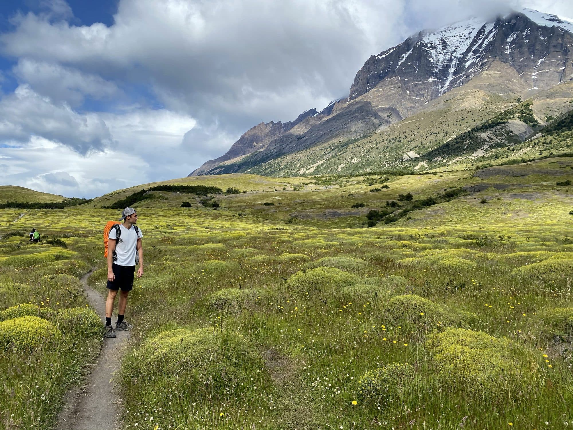 A hiker on a trail through a green valley below snowy peaks